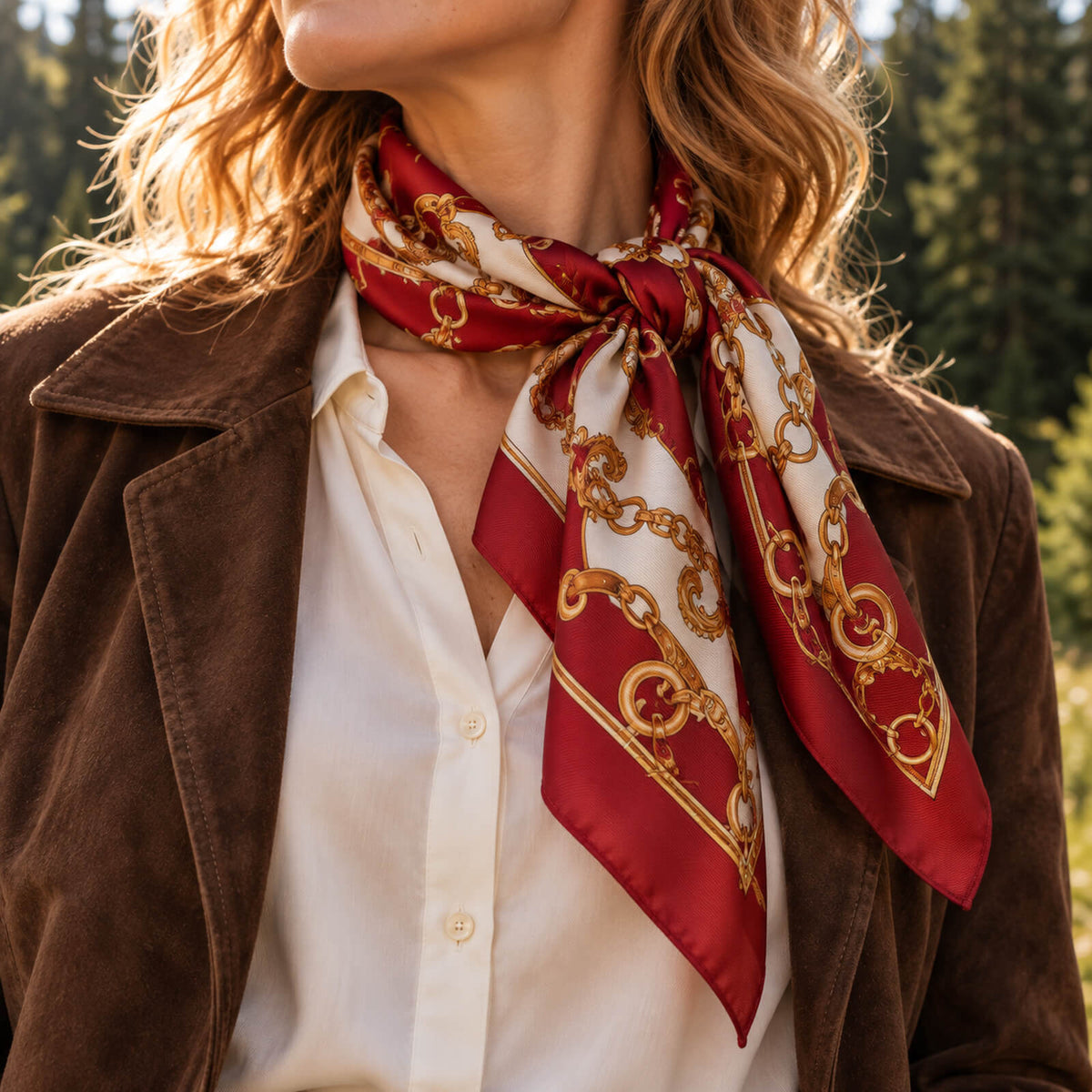 Woman wearing a brown jacket, white blouse, and a red silk scarf with gold chain patterns, standing outdoors with trees in the background.