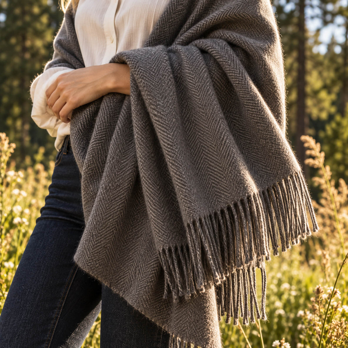 A person wearing a cream shirt and jeans stands outdoors, draped in a gray, fringed wool blanket. Sunlight shines on the field and tall grass around them, with trees blurred in the background.